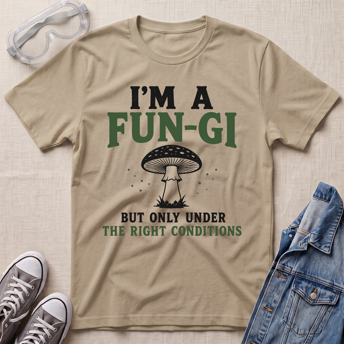 A beige graphic t-shirt laid flat on a textured fabric background, styled with safety goggles, gray sneakers, and a blue denim jacket. The t-shirt features a mushroom graphic and the text 'I'M A FUN-GI BUT ONLY UNDER THE RIGHT CONDITIONS'.