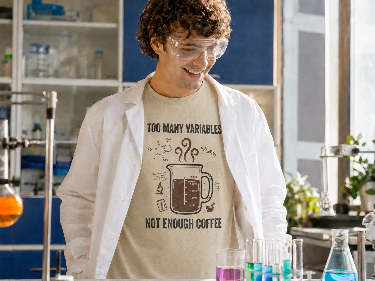 A man in a white lab coat and safety goggles wearing a beige graphic t-shirt that reads 'TOO MANY VARIABLES NOT ENOUGH COFFEE' with a coffee mug illustration, standing in a science lab.
