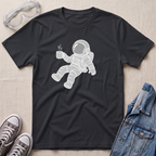 A black crewneck t-shirt featuring a white graphic of an astronaut floating and holding a steaming mug. The t-shirt is styled in a flat-lay arrangement on a textured beige background, surrounded by props including safety goggles, gray sneakers, and a denim jacket.