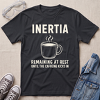 A black graphic t-shirt laid flat on a light beige textured background, surrounded by styling props including safety goggles, gray sneakers, and a denim jacket. The t-shirt features a white graphic of a steaming coffee mug with the text 'INERTIA REMAINING AT REST UNTIL THE CAFFEINE KICKS IN'.