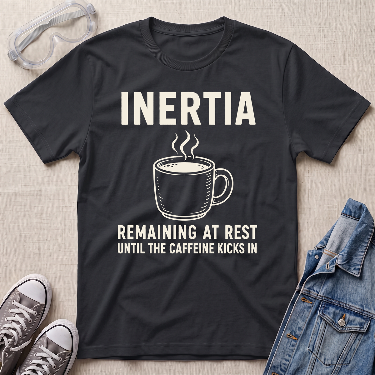 A black graphic t-shirt laid flat on a light beige textured background, surrounded by styling props including safety goggles, gray sneakers, and a denim jacket. The t-shirt features a white graphic of a steaming coffee mug with the text 'INERTIA REMAINING AT REST UNTIL THE CAFFEINE KICKS IN'.