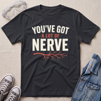 A black graphic t-shirt laid flat on a light beige textured background, surrounded by styling props including safety goggles, gray sneakers, and a denim jacket. The t-shirt features the text 'YOU'VE GOT A LOT OF NERVE' in white and orange lettering with an orange branch graphic underneath.