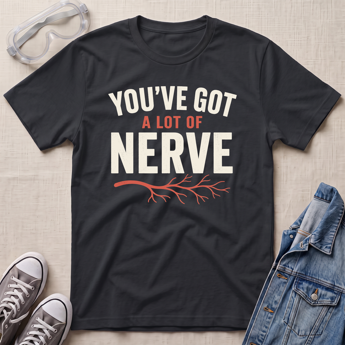 A black graphic t-shirt laid flat on a light beige textured background, surrounded by styling props including safety goggles, gray sneakers, and a denim jacket. The t-shirt features the text 'YOU'VE GOT A LOT OF NERVE' in white and orange lettering with an orange branch graphic underneath.