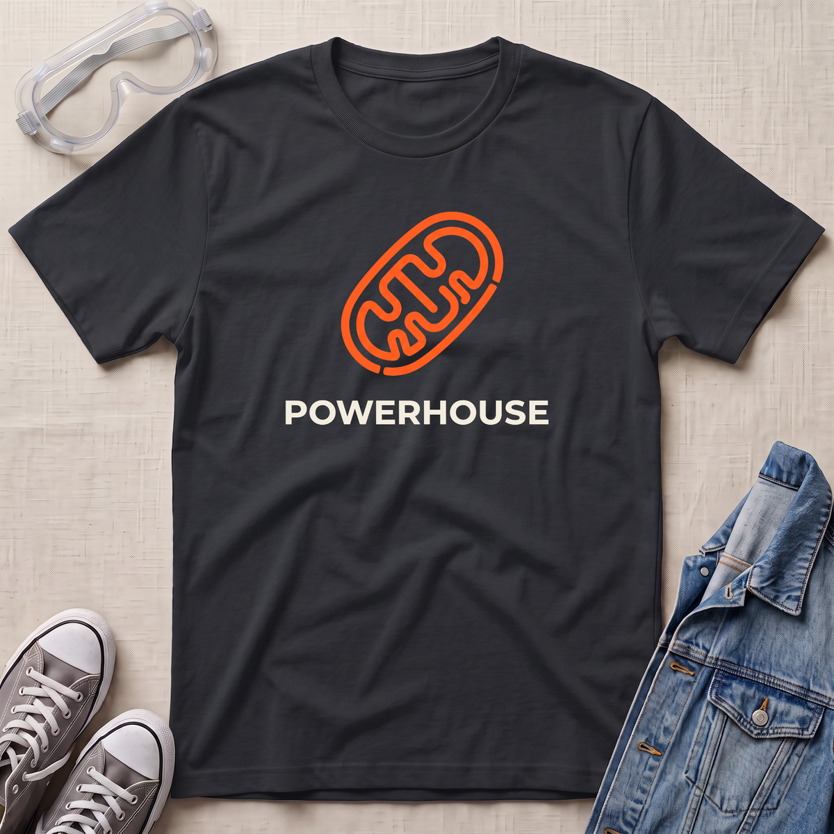 A black t-shirt with an orange mitochondrial outline and the word 'POWERHOUSE' in white, styled in a flat lay with jeans, sneakers, and safety goggles on a beige textured background.