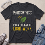 A dark charcoal gray graphic t-shirt laid flat on a beige textured background, surrounded by props including clear safety goggles, gray sneakers, and a blue denim jacket. The t-shirt features a green leaf graphic and the text 'PHOTOSYNTHESIS I'M A BIG FAN OF LIGHT WORK' in white and yellow.