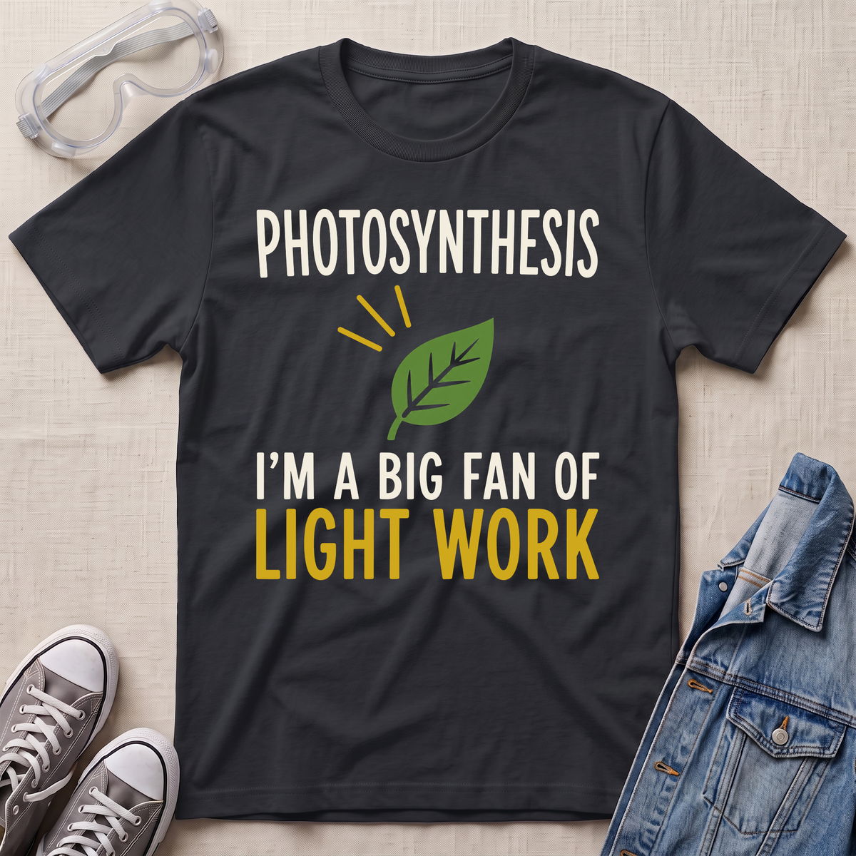 A dark charcoal gray graphic t-shirt laid flat on a beige textured background, surrounded by props including clear safety goggles, gray sneakers, and a blue denim jacket. The t-shirt features a green leaf graphic and the text 'PHOTOSYNTHESIS I'M A BIG FAN OF LIGHT WORK' in white and yellow.