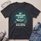 A black graphic t-shirt laid flat on a textured beige fabric background, styled with safety goggles, gray sneakers, and a denim jacket. The t-shirt features a circular graphic with a teal whale and the text 'MY KNOWLEDGE IS DEEP BUT MY HUMOR IS RATHER SHALLOW'.