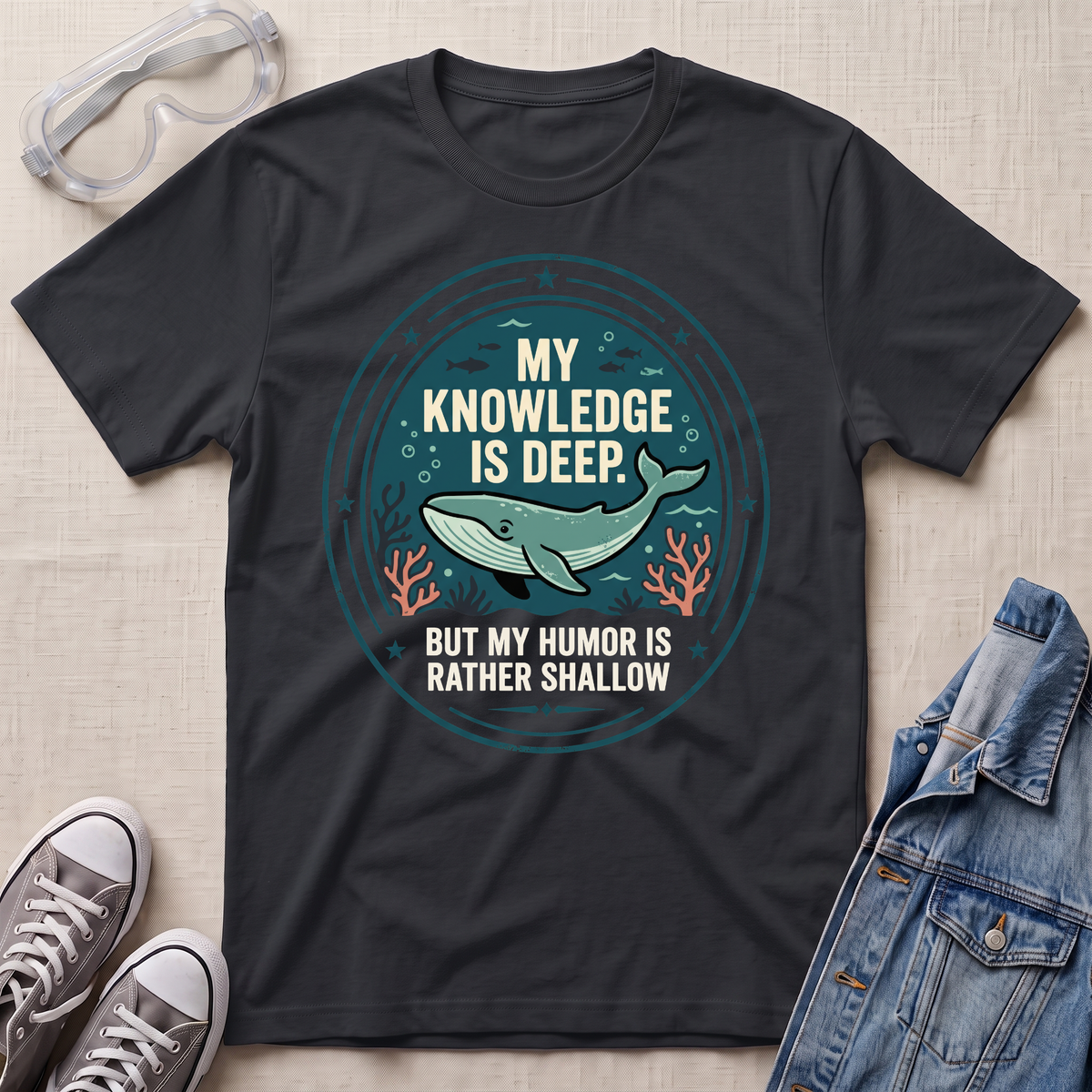 A black graphic t-shirt laid flat on a textured beige fabric background, styled with safety goggles, gray sneakers, and a denim jacket. The t-shirt features a circular graphic with a teal whale and the text 'MY KNOWLEDGE IS DEEP BUT MY HUMOR IS RATHER SHALLOW'.