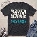 A black graphic t-shirt laid flat on a light beige textured background, surrounded by styling props including safety goggles, gray sneakers, and a denim jacket. The t-shirt features the text 'MY CHEMISTRY JOKES KEEP DISAPPEARING' in white and 'THEY ARGON' in bright teal blue.