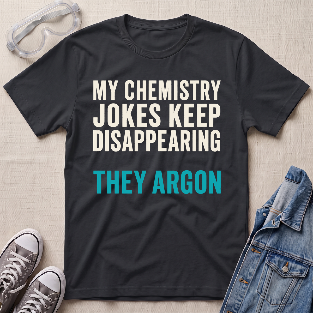 A black graphic t-shirt laid flat on a light beige textured background, surrounded by styling props including safety goggles, gray sneakers, and a denim jacket. The t-shirt features the text 'MY CHEMISTRY JOKES KEEP DISAPPEARING' in white and 'THEY ARGON' in bright teal blue.