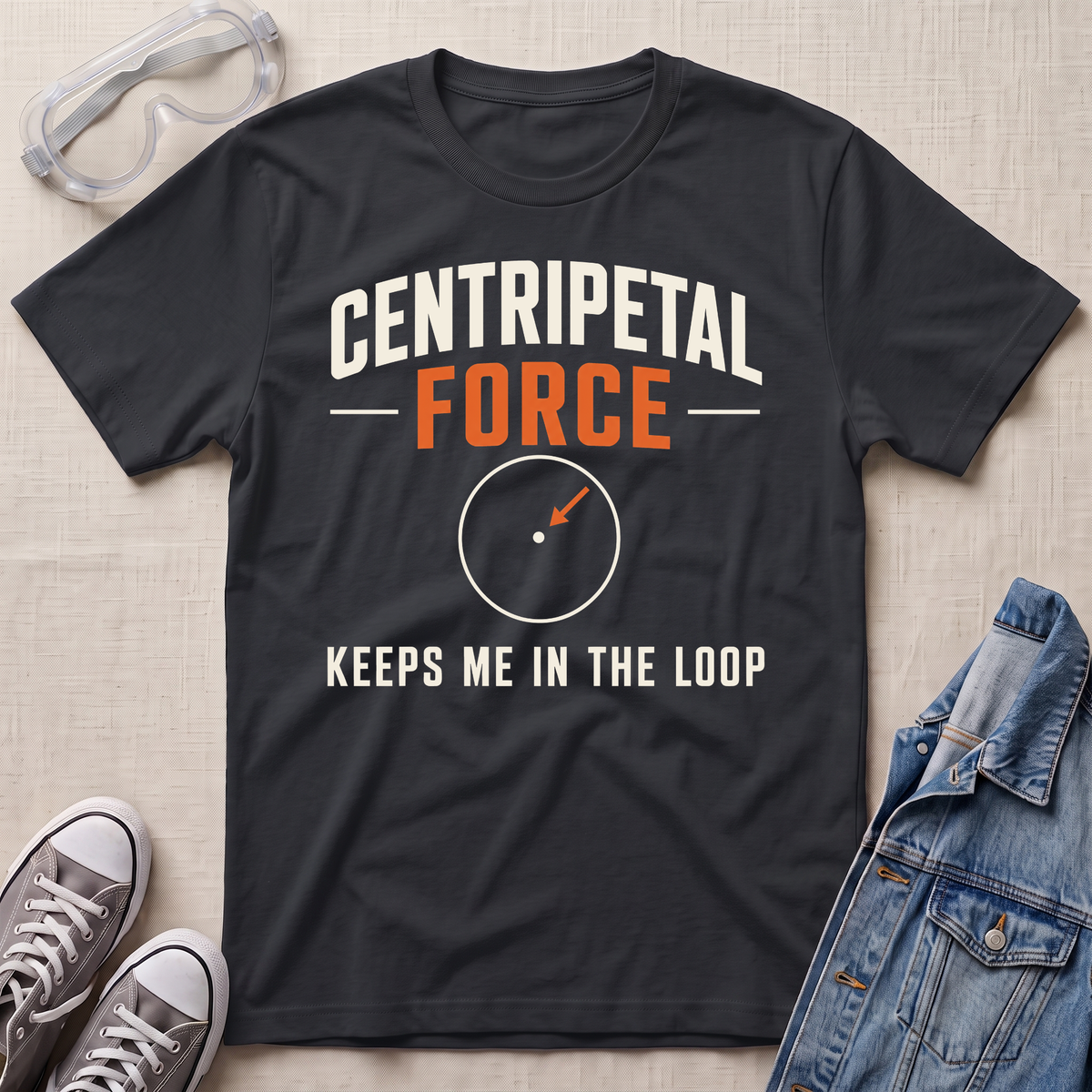 A black graphic t-shirt laid flat on a light beige textured background, styled with safety goggles, gray sneakers, and a denim jacket. The t-shirt features the text 'CENTRIPETAL FORCE KEEPS ME IN THE LOOP' in white and orange lettering with a small circular diagram graphic.