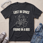 A black graphic t-shirt laid flat on a light beige fabric background, surrounded by styling props including clear safety goggles, gray sneakers, and a denim jacket. The t-shirt features a white line-art graphic of a cat in a space helmet popping out of a box, with the text 'LOST IN SPACE' above and 'FOUND IN A BOX' below.