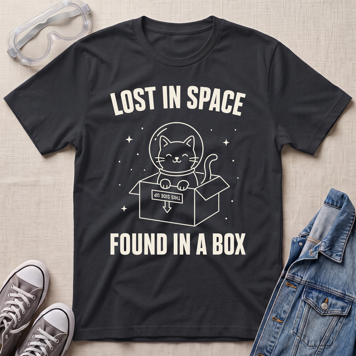 A black graphic t-shirt laid flat on a light beige fabric background, surrounded by styling props including clear safety goggles, gray sneakers, and a denim jacket. The t-shirt features a white line-art graphic of a cat in a space helmet popping out of a box, with the text 'LOST IN SPACE' above and 'FOUND IN A BOX' below.