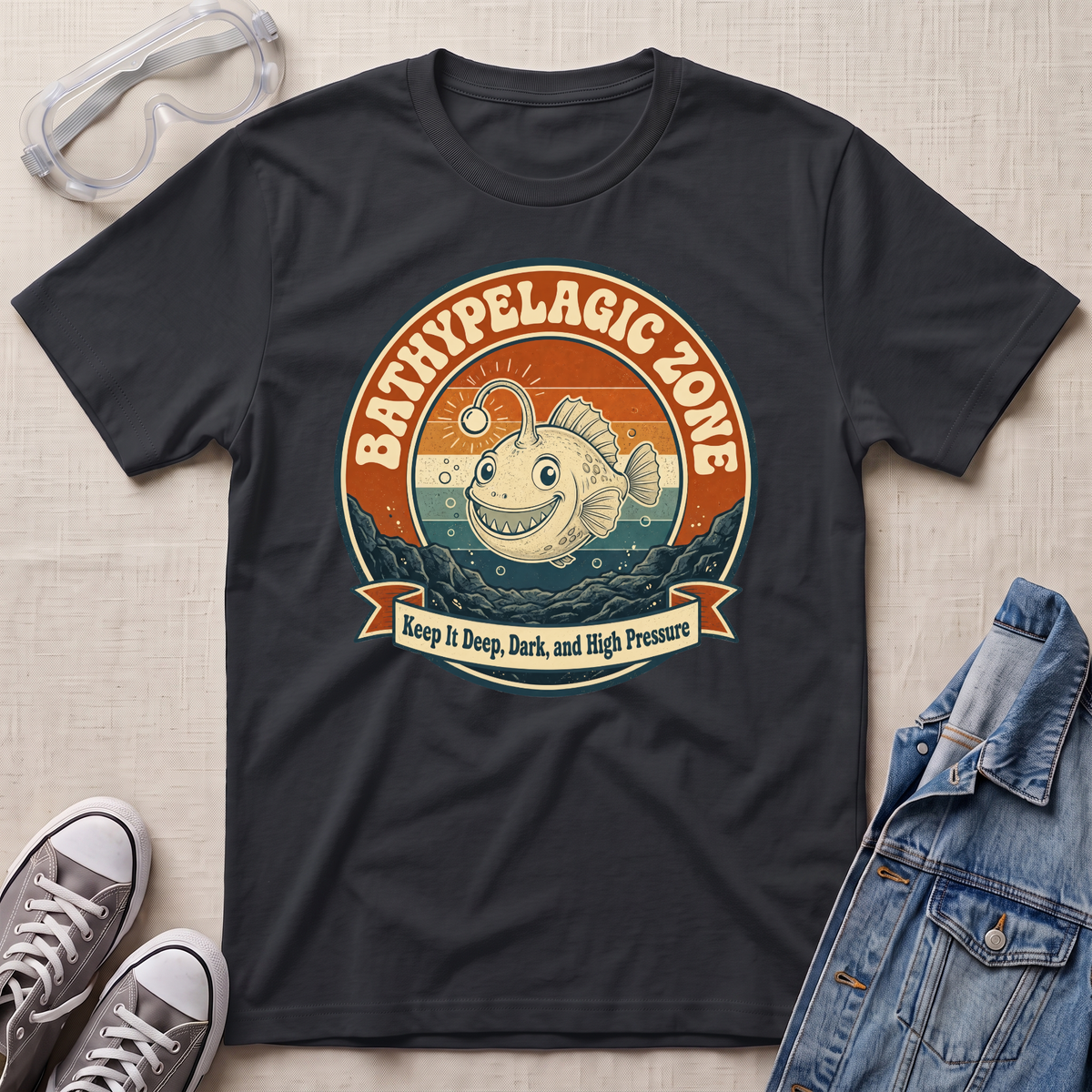 A black graphic t-shirt laid flat on a light beige textured background, styled with a pair of clear safety goggles, gray sneakers, and a blue denim jacket. The t-shirt features a retro-style circular graphic of a smiling fish in the bathypelagic zone with the text 'BATHYPELAGIC ZONE' and 'Keep it Deep, Dark, and High Pressure'.