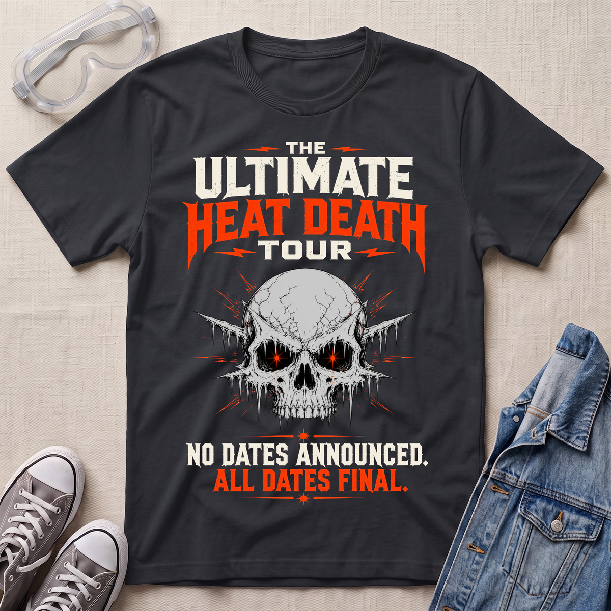 A black graphic t-shirt laid flat on a light beige textured background, styled with safety goggles, gray sneakers, and a denim jacket. The t-shirt features a skull graphic with glowing red eyes and bold text that reads 'THE ULTIMATE HEAT DEATH TOUR' and 'NO DATES ANNOUNCED.