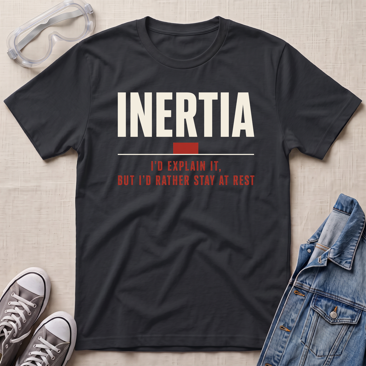 A black graphic t-shirt laid flat on a textured beige background, styled with safety goggles, gray sneakers, and a denim jacket. The t-shirt features the word 'INERTIA' in bold white letters, with a small red block and red text underneath reading 'I'D EXPLAIN IT, BUT I'D RATHER STAY AT REST'.