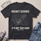 A black graphic t-shirt featuring white text and rocket science drawings, styled in a flat-lay with safety goggles, sneakers, and a denim jacket on a beige fabric background.