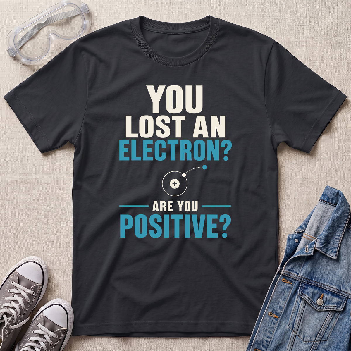 A black graphic t-shirt laid flat on a light beige textured background, surrounded by styled props including safety goggles, gray sneakers, and a denim jacket. The t-shirt features a white and light blue text graphic that reads 'YOU LOST AN ELECTRON? ARE YOU POSITIVE?' with a simple atom diagram.