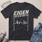 A black graphic t-shirt featuring the text 'EIGEN STAY IN MY OWN LANE' and the equation 'Av = λv' in distressed white print. The t-shirt is displayed in a flat-lay arrangement on a beige textured background, styled with safety goggles, gray sneakers, and a denim jacket.