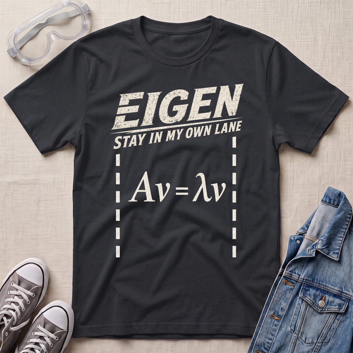 A black graphic t-shirt featuring the text 'EIGEN STAY IN MY OWN LANE' and the equation 'Av = λv' in distressed white print. The t-shirt is displayed in a flat-lay arrangement on a beige textured background, styled with safety goggles, gray sneakers, and a denim jacket.