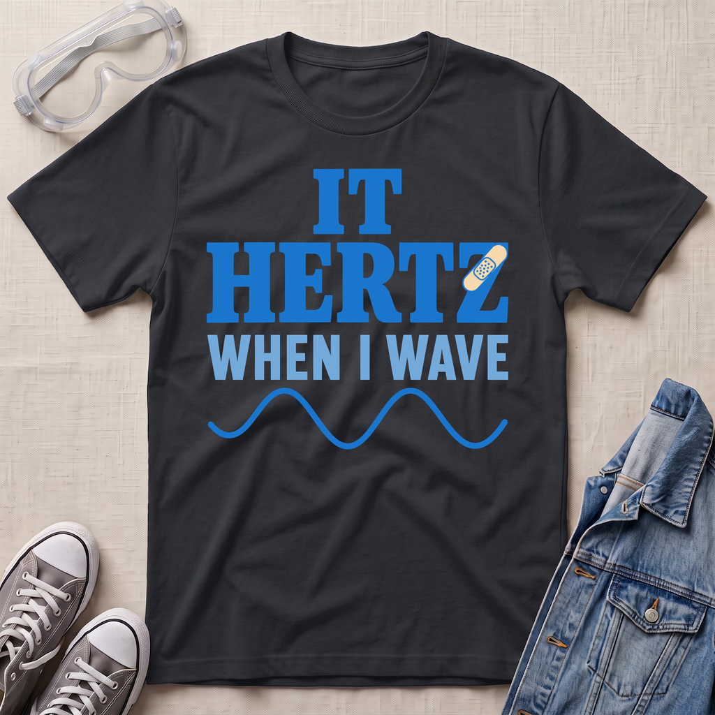A black graphic t-shirt laid flat on a light beige textured background, surrounded by styling props including safety goggles, gray sneakers, and a denim jacket. The t-shirt features the text 'IT HERTZ WHEN I WAVE' in blue and white, with a small band-aid graphic and blue wave line.