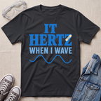 A black graphic t-shirt laid flat on a light beige textured background, surrounded by styling props including safety goggles, gray sneakers, and a denim jacket. The t-shirt features the text 'IT HERTZ WHEN I WAVE' in blue and white, with a small band-aid graphic and blue wave line.