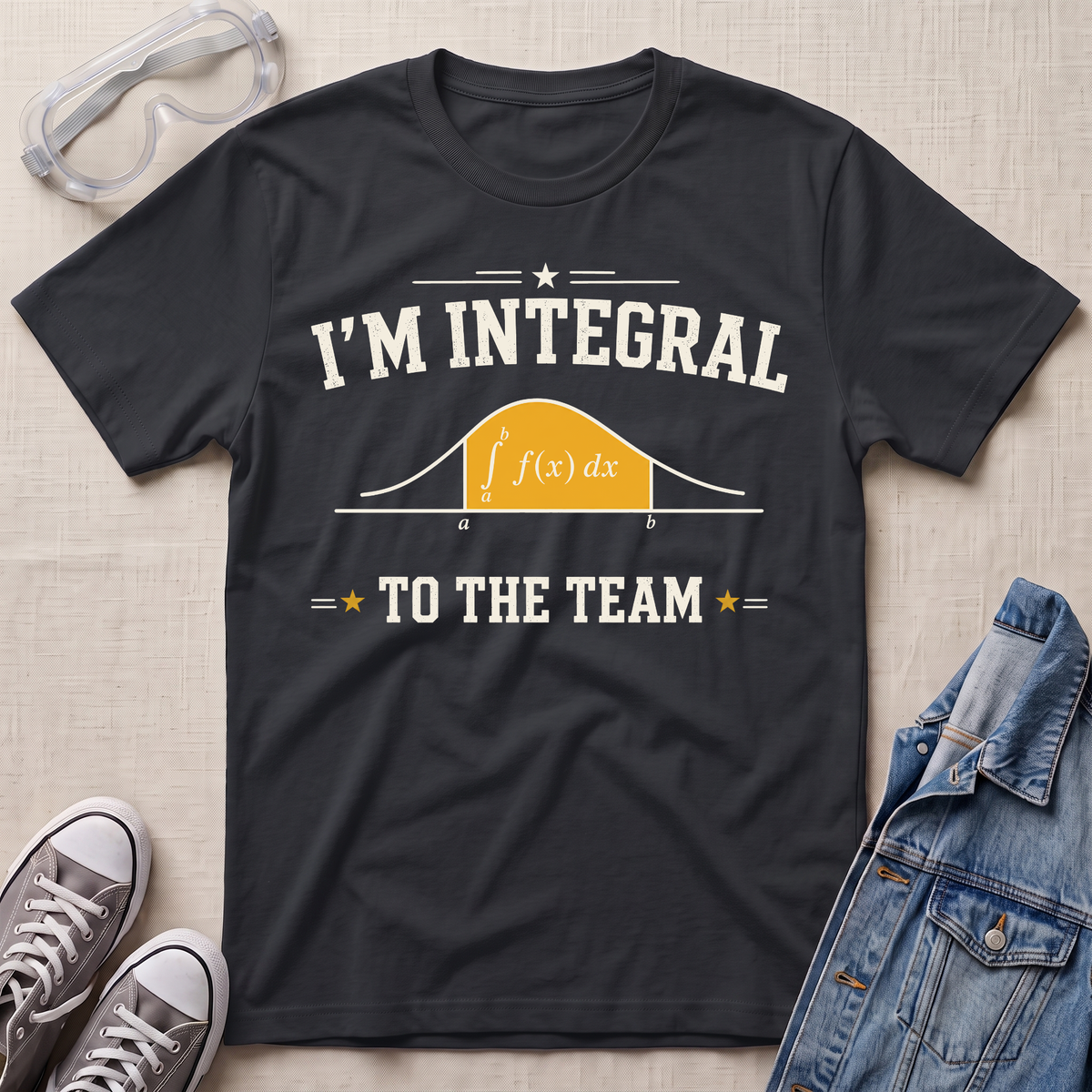 A navy blue graphic t-shirt laid flat on a beige fabric background, styled with safety goggles, gray sneakers, and a denim jacket. The t-shirt features a white graphic that reads 'I'M INTEGRAL TO THE TEAM' with a yellow integral curve diagram.