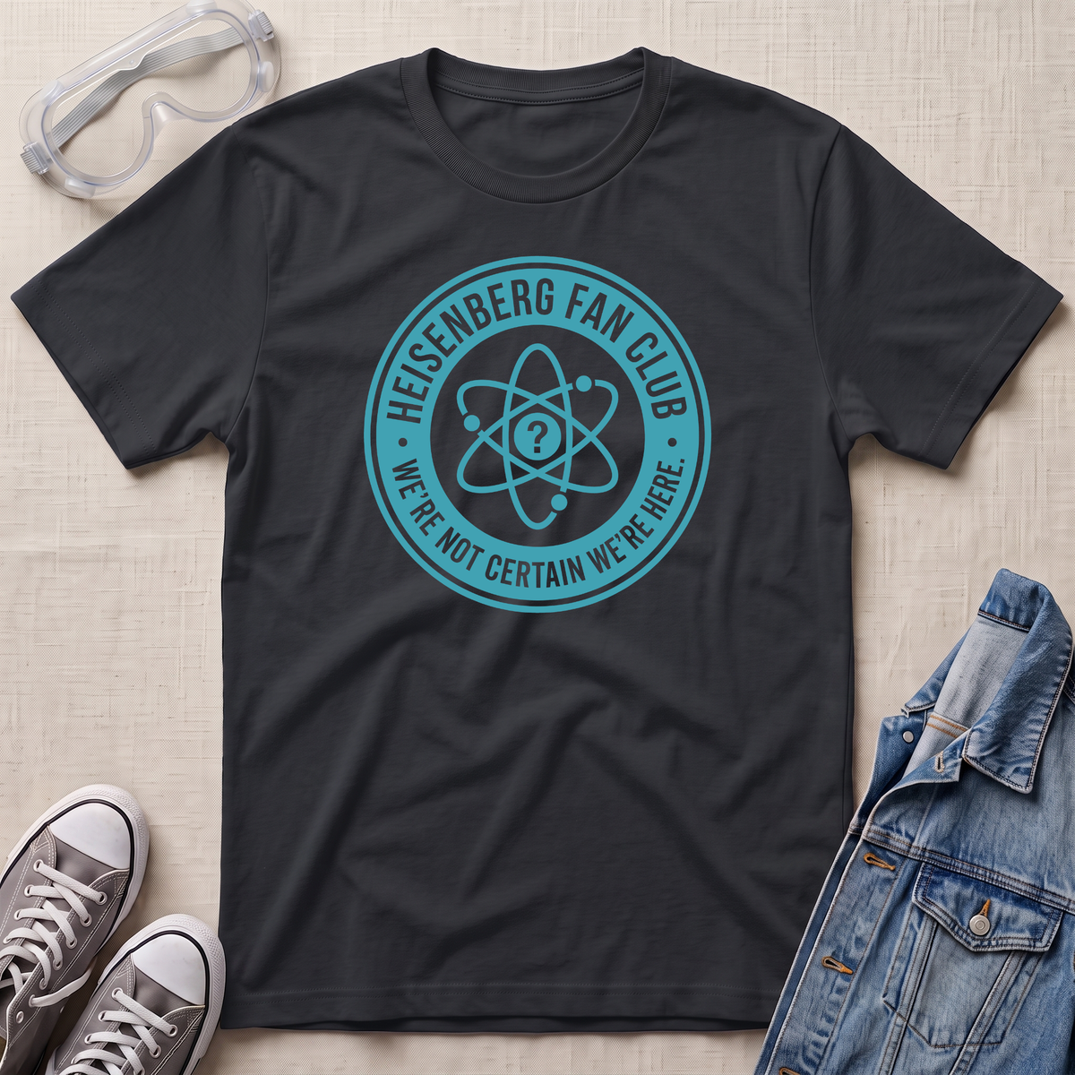 A black t-shirt laid flat on a light beige textured background, featuring a teal circular Heisenberg fan club graphic with an atom symbol and the text 'HEISENBERG FAN CLUB' and 'WE'RE NOT CERTAIN WE'RE HERE.' Surrounding props include safety goggles, gray sneakers, and a denim jacket.