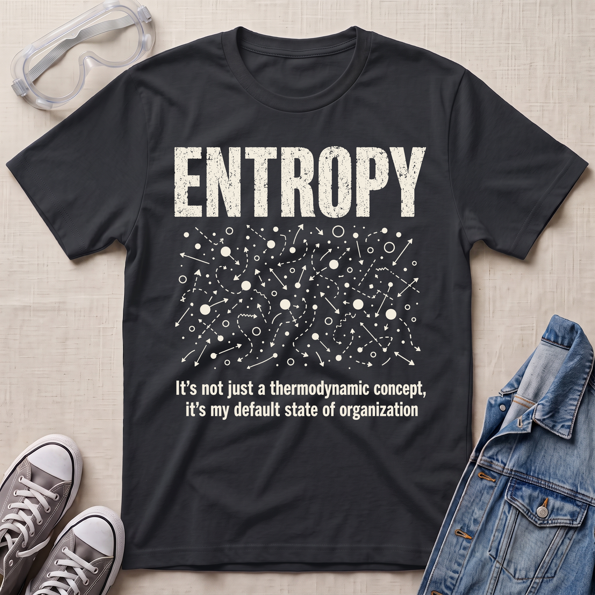 A black graphic t-shirt laid flat on a beige fabric background, styled with safety goggles, gray sneakers, and a denim jacket. The t-shirt features a white distressed graphic of the word 'ENTROPY' above a entropy pattern, with the text 'It's not just a thermodynamic concept, it's my default state of organization' below.