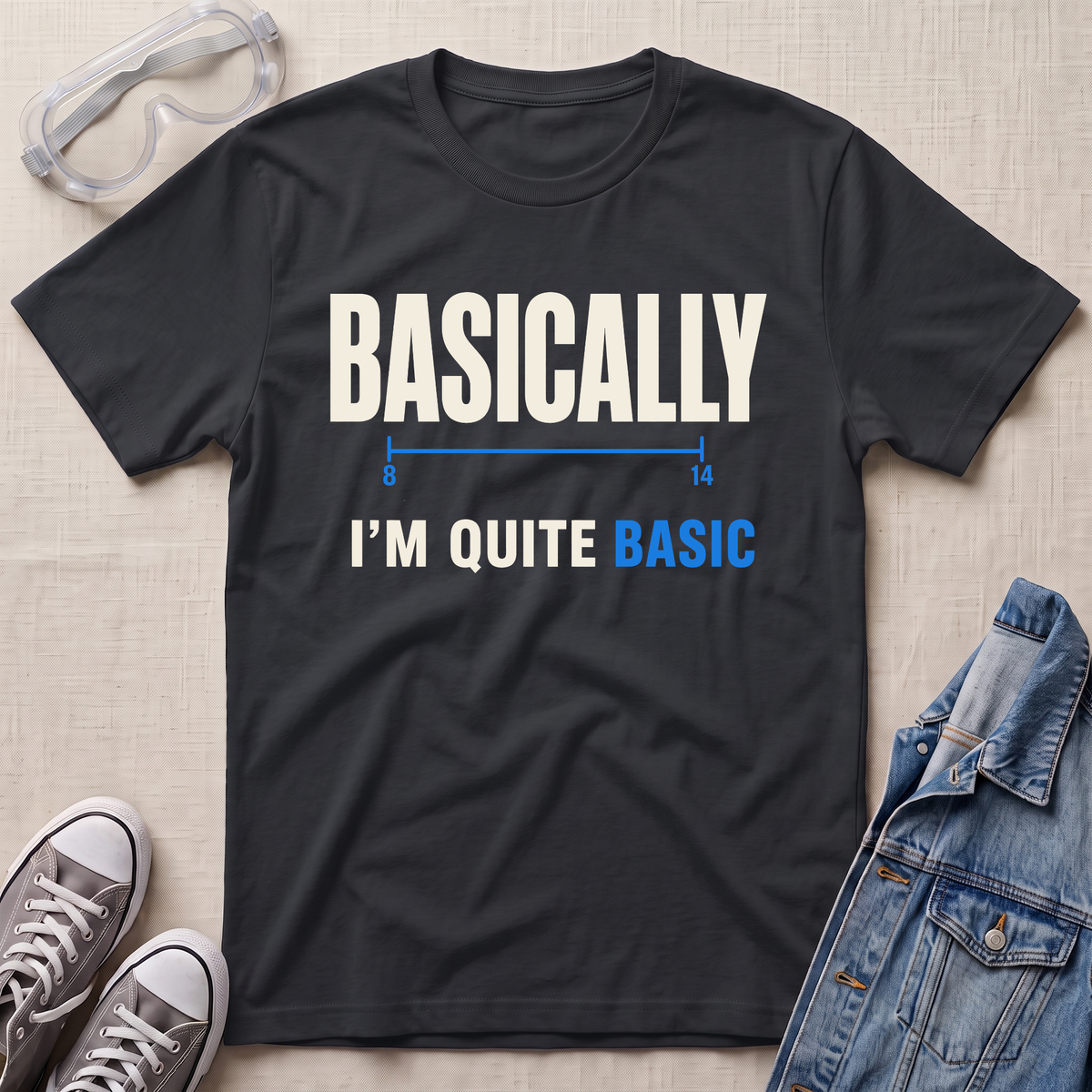A black graphic t-shirt laid flat on a light beige textured background, styled with safety goggles, gray sneakers, and a denim jacket. The t-shirt features the text 'BASICALLY I'M QUITE BASIC' in white and blue lettering.