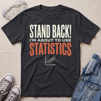 A black graphic t-shirt laid flat on a beige fabric background, surrounded by props including safety goggles, gray sneakers, and a denim jacket. The t-shirt features the text 'STAND BACK! I'M ABOUT TO USE STATISTICS' in white and orange lettering with a small scatterplot graphic below.