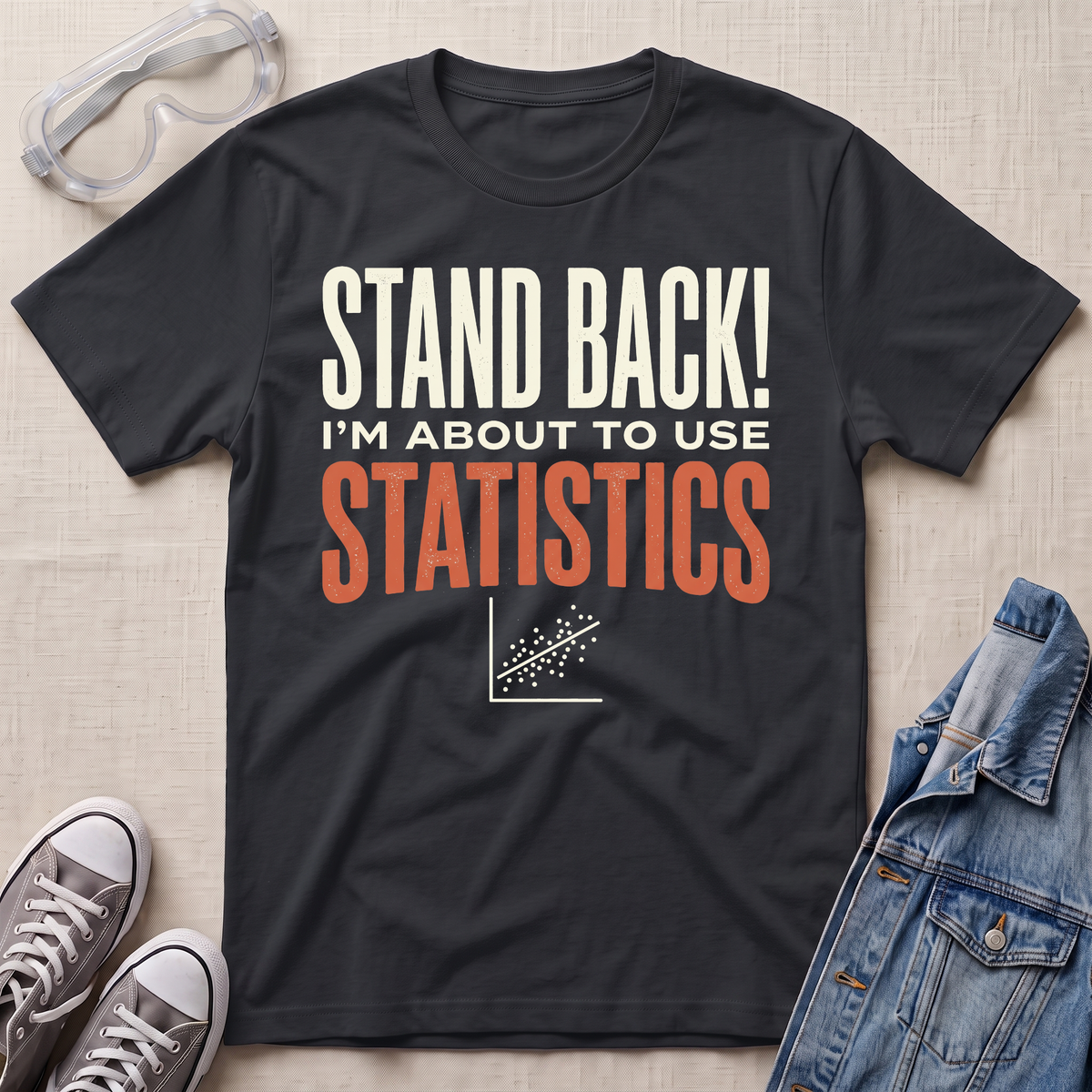 A black graphic t-shirt laid flat on a beige fabric background, surrounded by props including safety goggles, gray sneakers, and a denim jacket. The t-shirt features the text 'STAND BACK! I'M ABOUT TO USE STATISTICS' in white and orange lettering with a small scatterplot graphic below.