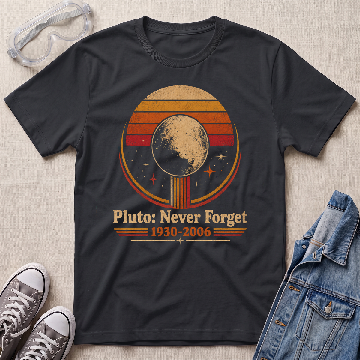 A black graphic t-shirt laid flat on a light beige textured background, styled with clear safety goggles, gray sneakers, and a blue denim jacket. The t-shirt features a retro circular graphic of Pluto with the text 'Pluto: Never Forget 1930-2006'.