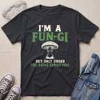 A black graphic t-shirt laid flat on a light beige textured background, styled with safety goggles, gray sneakers, and a denim jacket. The t-shirt features a white and green graphic of a mushroom with the text 'I'M A FUN-GI BUT ONLY UNDER THE RIGHT CONDITIONS'.