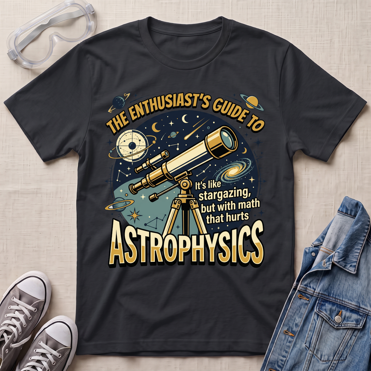 A black graphic t-shirt laid flat on a light beige textured background, styled with clear safety goggles, gray sneakers, and a blue denim jacket. The t-shirt features a detailed graphic of a telescope, planets, stars, and text that reads 'THE ENTHUSIAST'S GUIDE TO ASTROPHYSICS'.