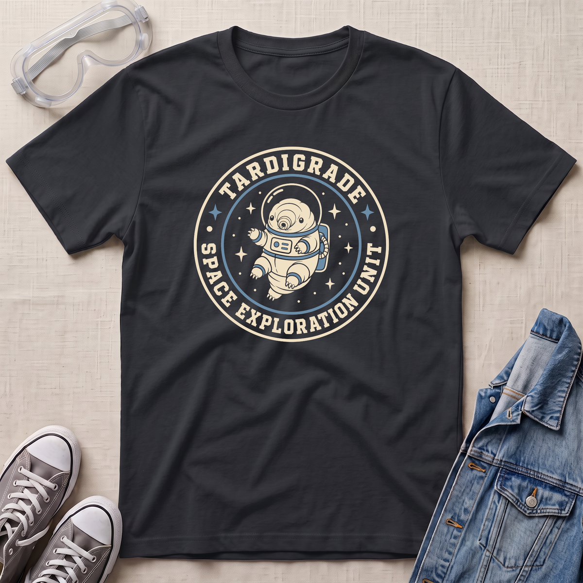 A black graphic t-shirt laid flat on a light beige textured background, styled with a pair of clear safety goggles, gray sneakers, and a blue denim jacket. The t-shirt features a circular graphic of an astronaut floating in space with the text 'TARDIGRADE SPACE EXPLORATION UNIT'.