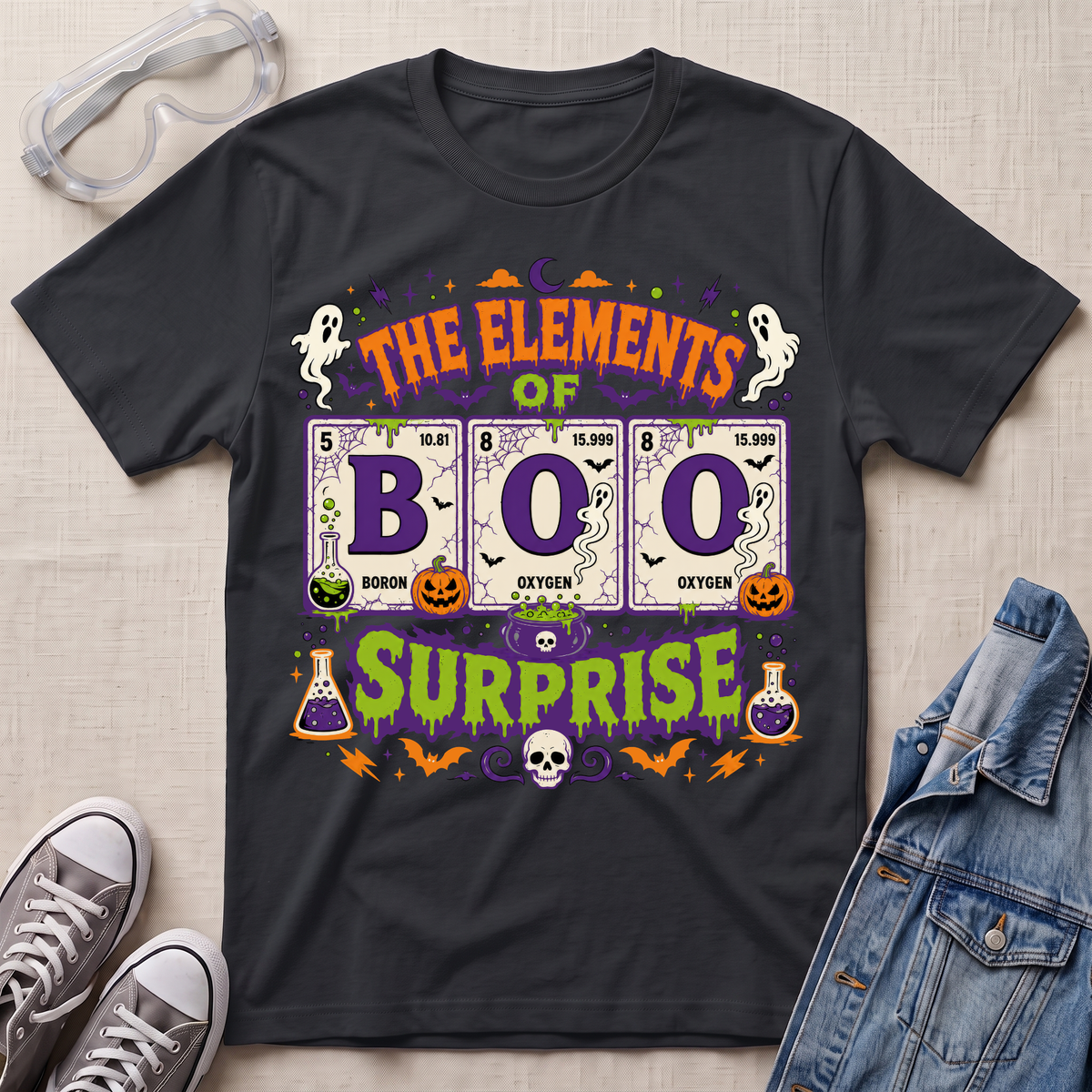 A black graphic t-shirt featuring a Halloween-themed design with the text 'THE ELEMENTS OF BOO SURPRISE' and periodic table elements styled as potion bottles, ghosts, pumpkins, and a skull. The shirt is displayed in a flat-lay arrangement with safety goggles, gray sneakers, and a denim jacket on a light beige textured background.