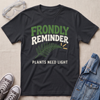 A black graphic t-shirt laid flat on a light beige textured background, styled with safety goggles, gray sneakers, and a denim jacket. The t-shirt features a green fern leaf graphic and the text 'FRONDLY REMINDER PLANTS NEED LIGHT' in green and white.