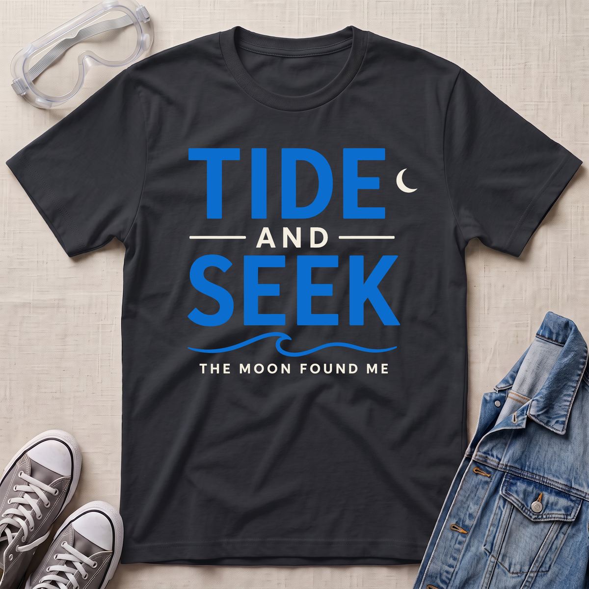 A black short-sleeve graphic T-shirt laid flat on a light beige textured surface, with safety glasses, gray sneakers, and a folded denim jacket around it. The shirt has large navy text reading 'TIDE AND SEEK' with smaller text 'THE MOON FOUND ME' and a crescent moon graphic.