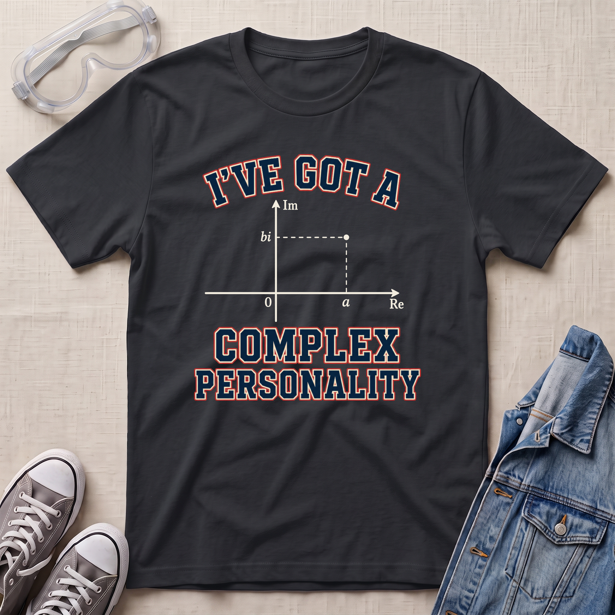 A black graphic t-shirt laid flat on a textured beige fabric background, styled with safety goggles, gray sneakers, and a denim jacket. The t-shirt features a graphic of a complex number plot with the text 'I'VE GOT A COMPLEX PERSONALITY' in red and blue outlined lettering.