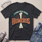 A black graphic t-shirt laid flat on a light beige textured background, surrounded by styling props including clear safety goggles, gray sneakers, and a blue denim jacket. The t-shirt features a graphic of a bone with the text 'I FIND THIS SHIRT HUMERUS' in stylized orange and teal lettering.