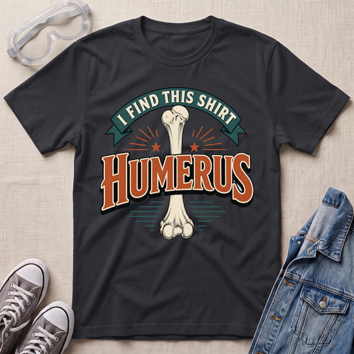 A black graphic t-shirt laid flat on a light beige textured background, surrounded by styling props including clear safety goggles, gray sneakers, and a blue denim jacket. The t-shirt features a graphic of a bone with the text 'I FIND THIS SHIRT HUMERUS' in stylized orange and teal lettering.
