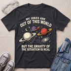 A black graphic t-shirt laid flat on a light beige textured surface, surrounded by styling props including clear safety glasses, gray sneakers, and a blue denim jacket. The t-shirt features a space-themed graphic with planets, stars, and the text 'MY JOKES ARE OUT OF THIS WORLD BUT THE GRAVITY OF THE SITUATION IS REAL'.