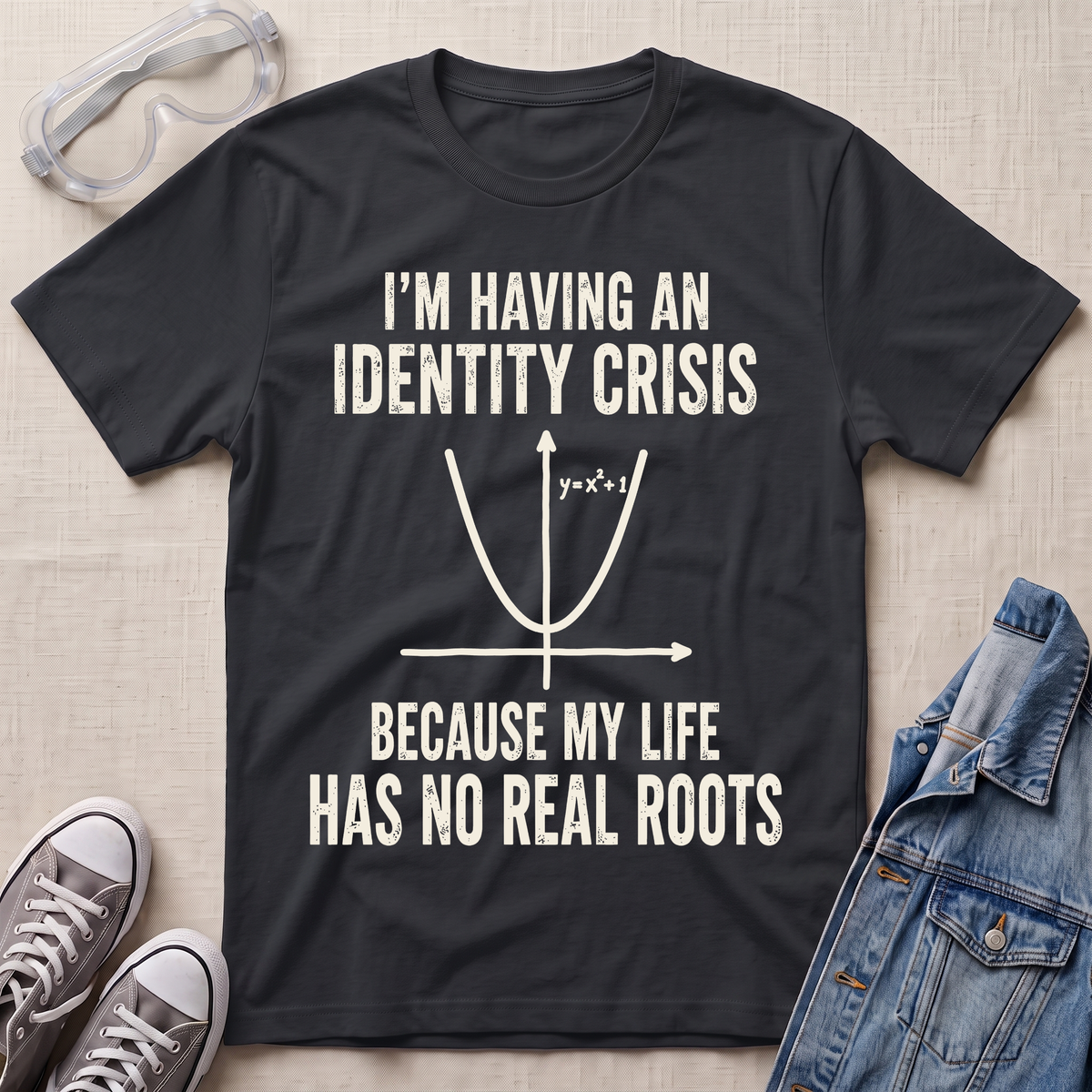 A black graphic t-shirt laid flat on a beige fabric background, surrounded by styling props including clear safety goggles, gray sneakers, and a denim jacket. The t-shirt features a white printed graphic of a parabola with the text 'I'M HAVING AN IDENTITY CRISIS BECAUSE MY LIFE HAS NO REAL ROOTS'.