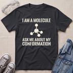 A black graphic t-shirt laid flat on a light beige textured background, surrounded by styling props including safety goggles, gray sneakers, and a denim jacket. The t-shirt features a white printed text design that reads 'I AM A MOLECULE ASK ME ABOUT MY CONFORMATION' with a simple molecular model graphic.