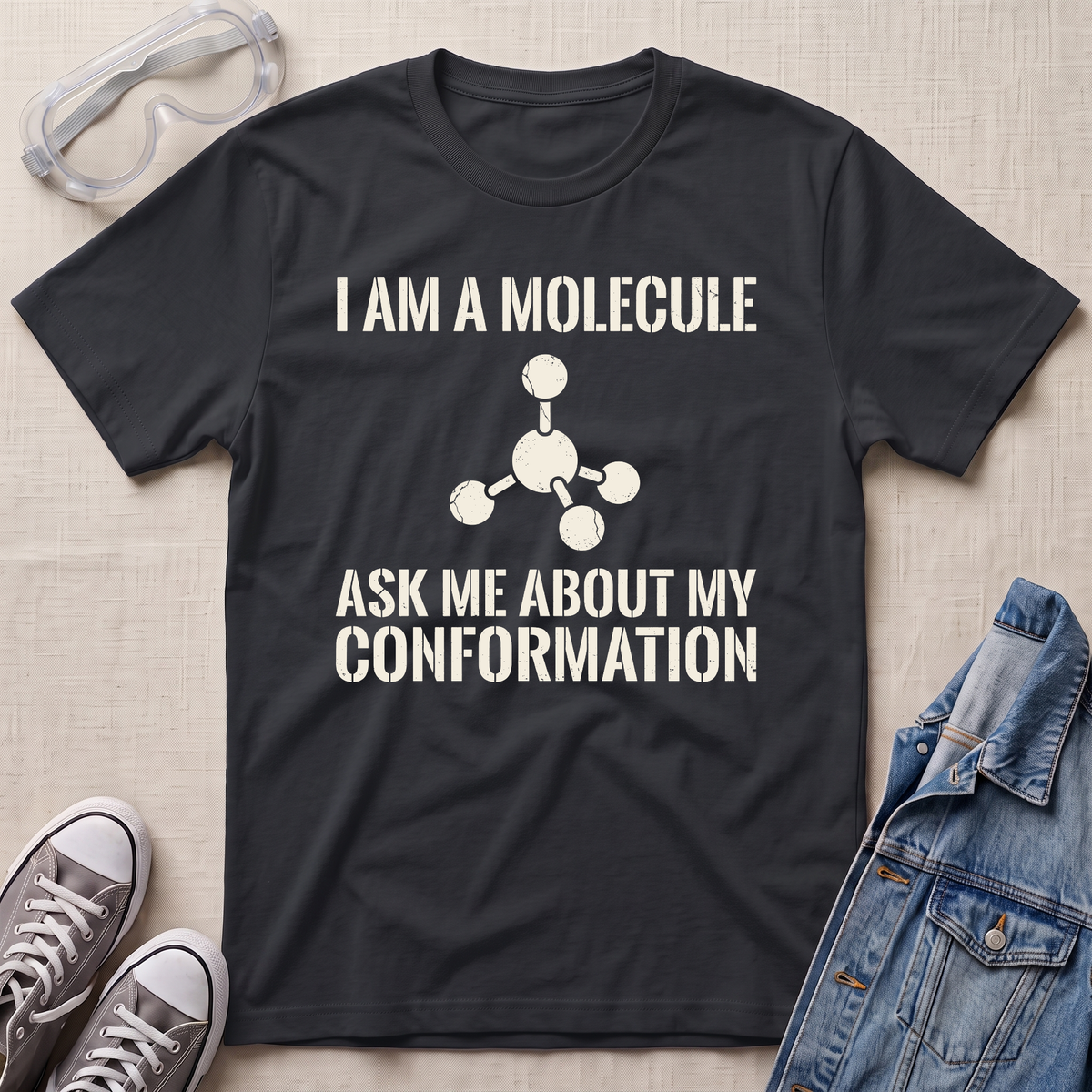 A black graphic t-shirt laid flat on a light beige textured background, surrounded by styling props including safety goggles, gray sneakers, and a denim jacket. The t-shirt features a white printed text design that reads 'I AM A MOLECULE ASK ME ABOUT MY CONFORMATION' with a simple molecular model graphic.
