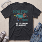 A black graphic t-shirt laid flat on a light beige fabric background, styled with safety goggles, gray sneakers, and a denim jacket. The t-shirt features a colorful retro toy gun illustration with the text 'PEW! PEW! PEW! ... IS THE SOUND OF SCIENCE.'