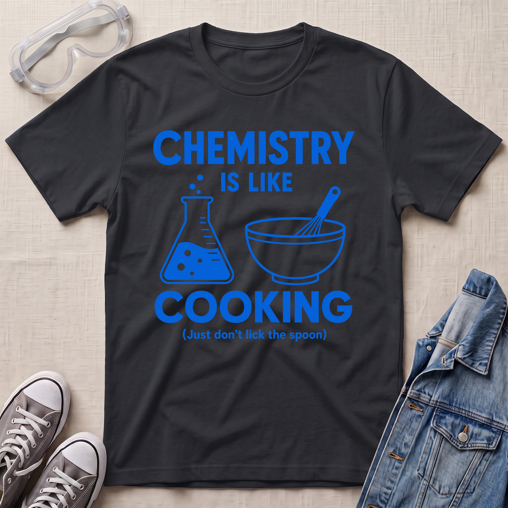 A black graphic t-shirt laid flat on a beige textured background, surrounded by styling props including safety goggles, gray sneakers, and a denim jacket. The t-shirt features a blue graphic that reads 'CHEMISTRY IS LIKE COOKING (Just don't lick the spoon)' with illustrations of a beaker and a mixing bowl.