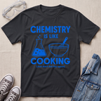 A black graphic t-shirt laid flat on a beige textured background, surrounded by styling props including safety goggles, gray sneakers, and a denim jacket. The t-shirt features a blue graphic that reads 'CHEMISTRY IS LIKE COOKING (Just don't lick the spoon)' with illustrations of a beaker and a mixing bowl.
