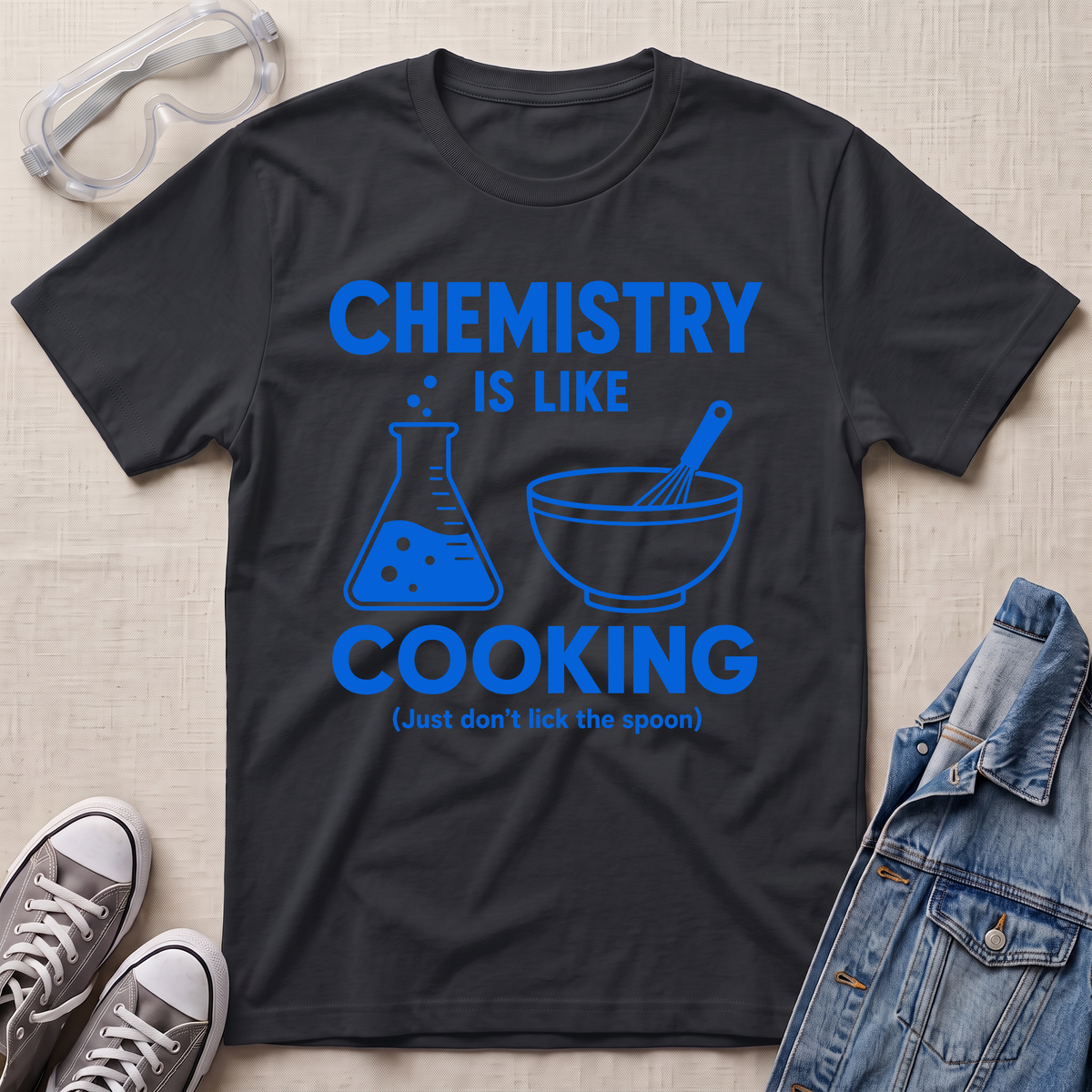 A black graphic t-shirt laid flat on a beige textured background, surrounded by styling props including safety goggles, gray sneakers, and a denim jacket. The t-shirt features a blue graphic that reads 'CHEMISTRY IS LIKE COOKING (Just don't lick the spoon)' with illustrations of a beaker and a mixing bowl.